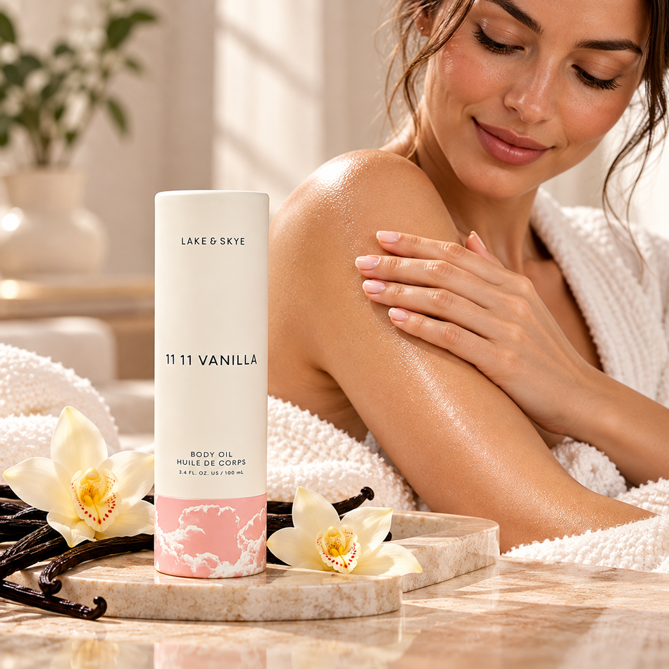 Lake & Skye 11 11 Vanilla Body Oil 3.4 oz (100 ml) displayed in a luxury spa setting with a woman applying body oil to her shoulder, surrounded by vanilla flowers and soft towels.