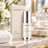 SkinMedica TNS Eye Repair 0.5 oz (15 mL) eye cream displayed with matching ivory packaging, white flowers, and a soft towel on a luxury marble skincare vanity.